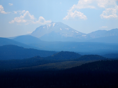 [From near to far the hills become taller and more grey. Lassen Peak is in the far distance with triangles of snow coming down from the top in several places. The near hills are covered in evergreens.]